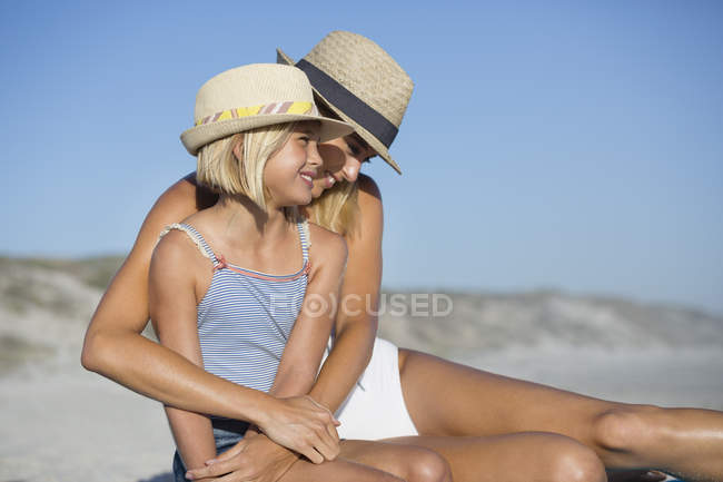 Smiling woman and daughter in hats sitting on sandy beach — Stock Photo