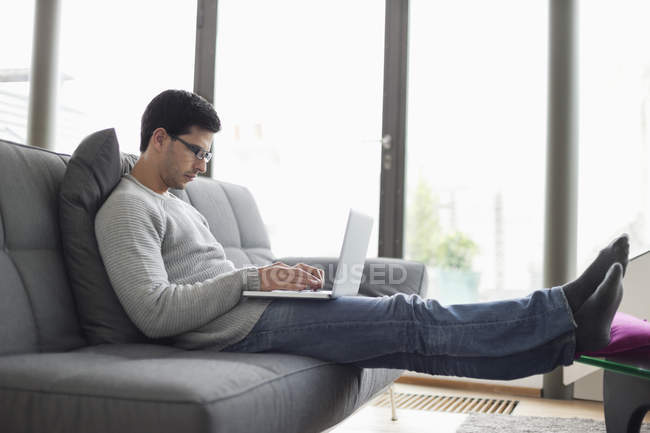 Hombre seguro de sí mismo usando el ordenador portátil en el sofá en casa - foto de stock