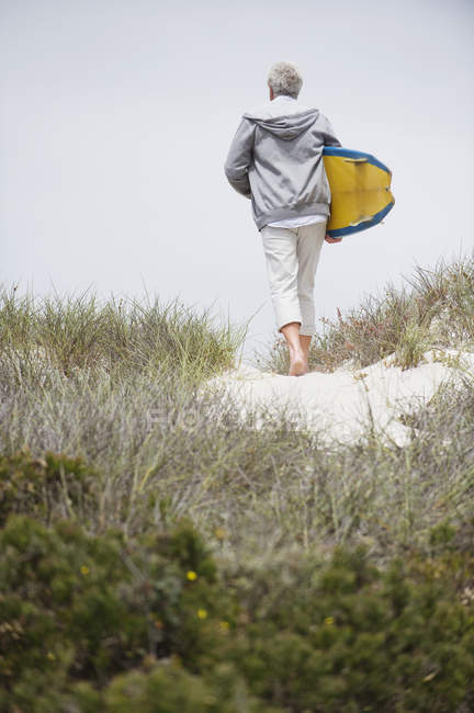 Вид сзади на старшего человека, несущего доску для серфинга на пляже — стоковое фото