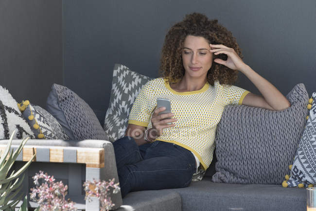 Жінка сидить на дивані вдома і використовує смартфон — стокове фото