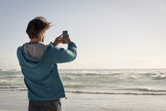Happy young man taking picture with mobile phone on beach — Stock Photo