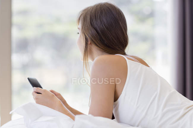 Junge Frau mit Handy auf Bett neben Fenster liegend — Stockfoto