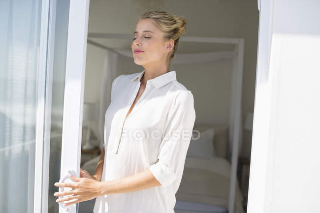 Jovem mulher relaxada com os olhos fechados em pé na janela à luz do sol — Fotografia de Stock
