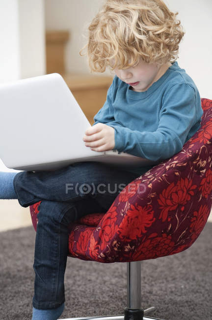 Милый мальчик с светлыми волосами с помощью ноутбука в кресле дома — стоковое фото
