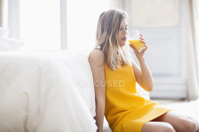 Woman in bright yellow dress drinking orange juice on floor at home — Stock Photo