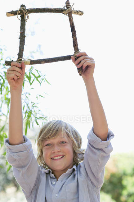 Portrait de mignon garçon tenant cadre de bois flotté avec les bras levés à l'extérieur — Photo de stock