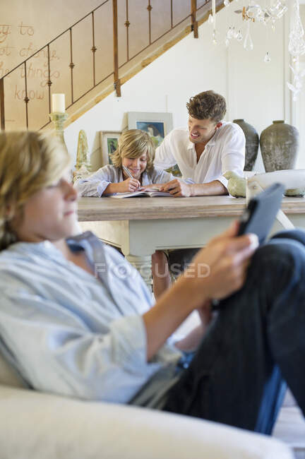 Человек обучает маленького мальчика с братом, используя цифровые таблетки на переднем плане в доме — стоковое фото
