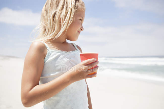Petite fille tenant tasse jetable sur la plage et regardant la vue — Photo de stock