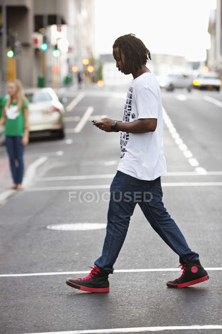 Mensajería de texto hombre joven con teléfono móvil mientras cruza la carretera - foto de stock