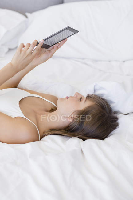 Молода жінка лежить на ліжку і читає електронну книгу — стокове фото