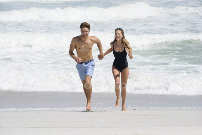 Счастливая пара в купальниках бегает по пляжу, держа за руки — стоковое фото