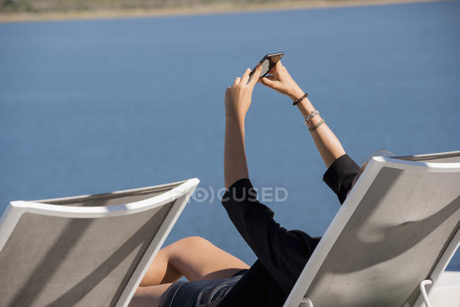 Mujer tomando selfie con el teléfono móvil en la silla de cubierta en el paseo marítimo - foto de stock