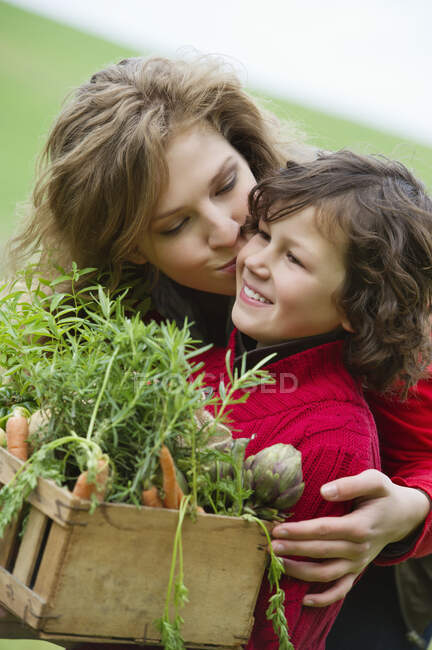 Menino segurando uma caixa de legumes com sua mãe beijando-o — Fotografia de Stock