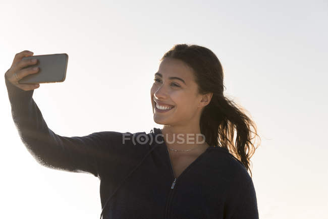 Mulher feliz tirando selfie com smartphone contra céu limpo — Fotografia de Stock