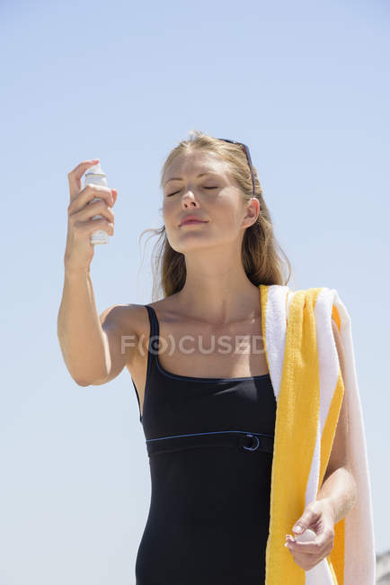 Mulher elegante em maiô pulverizando protetor solar no rosto contra o céu azul — Fotografia de Stock
