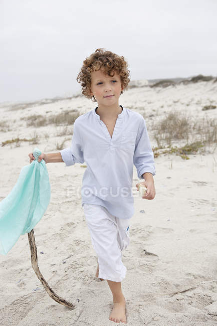 Мальчик держит флаг на палочке и ходит по песчаному пляжу — стоковое фото