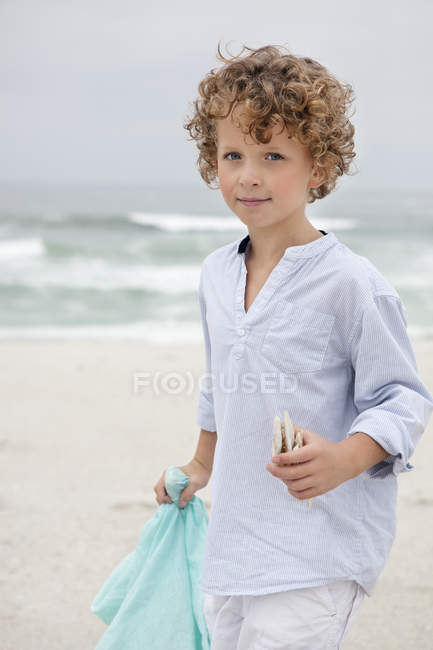 Porträt eines niedlichen Jungen, der mit Muscheln am Strand steht — Stockfoto