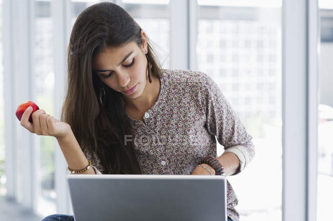 Focused woman holding apple while using a laptop — Stock Photo