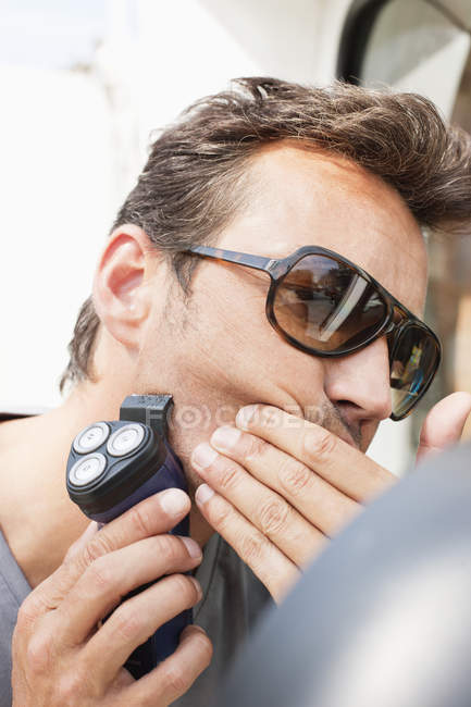 Чоловік в сонцезахисних окулярах використовує електричну бритву на обличчі — стокове фото