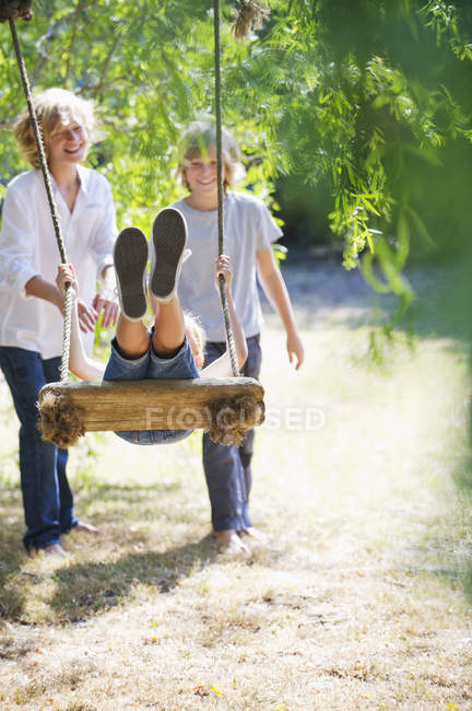 Усміхнені брати і сестри грають в гойдалки на дереві в літньому саду — стокове фото