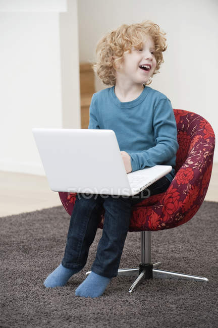 Cheerful boy with blonde hair using a laptop in armchair at home — Stock Photo
