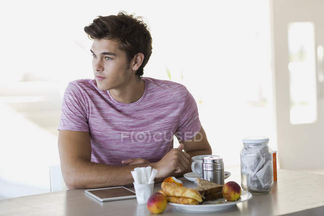 Молодой человек сидит за кухонным столом с чашкой кофе — стоковое фото