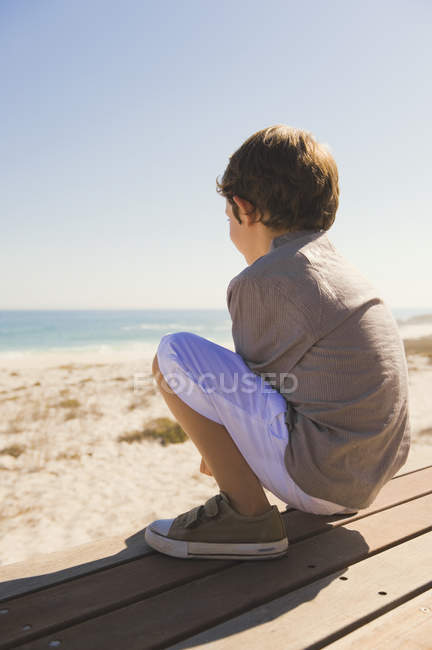Menino sentado no calçadão de madeira na costa marítima — Fotografia de Stock