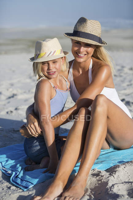Sonriente mujer e hija en sombreros sentado en la playa de arena - foto de stock