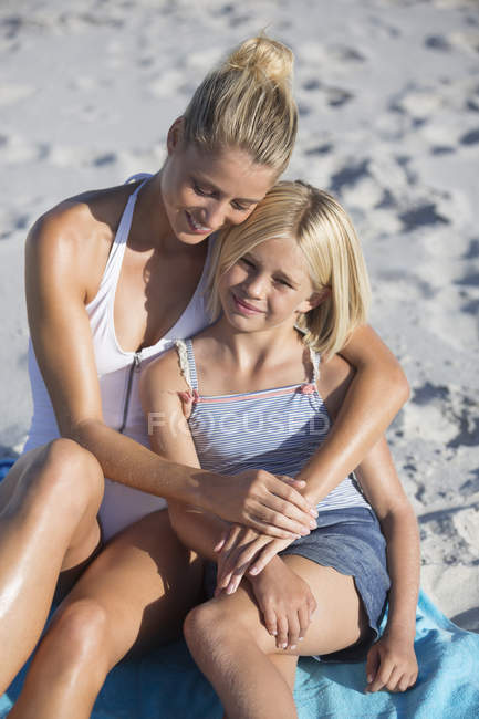 Feliz mujer e hija sentadas y abrazadas en la playa - foto de stock