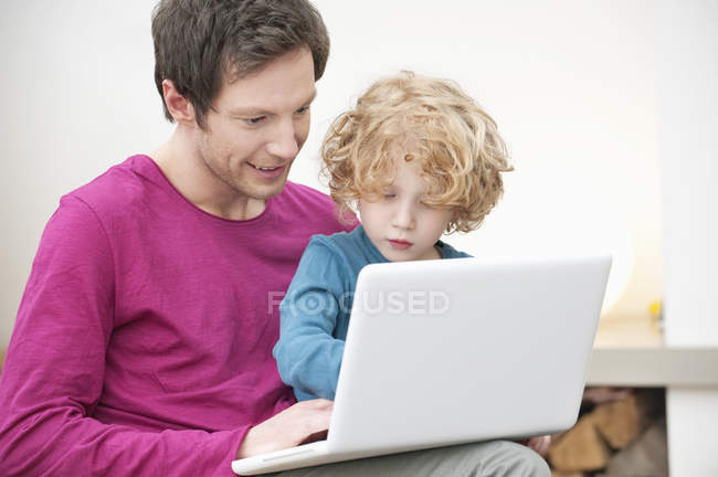 Чоловік допомагає маленькому синові використовувати ноутбук на дивані вдома — стокове фото