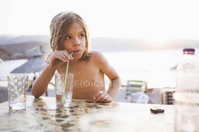 Niño con el pelo largo bebiendo a orillas del lago y mirando hacia otro lado - foto de stock