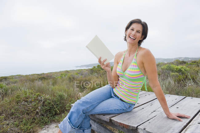 Laughing woman sitting on boardwalk in nature with book — Stock Photo