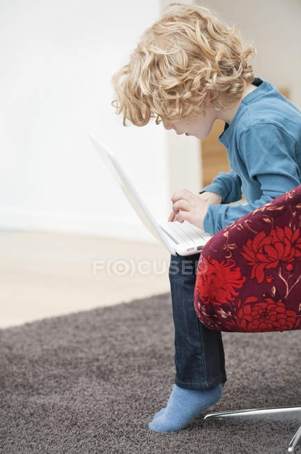 Cute boy with blonde hair using a laptop in armchair at home — Stock Photo