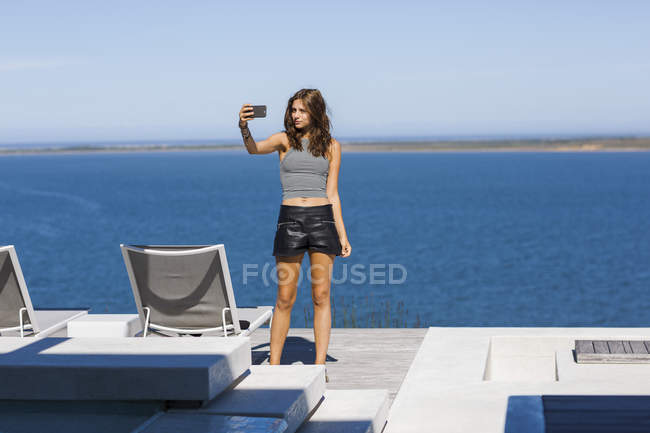 Stylische junge Frau macht Selfie mit Smartphone auf Terrasse am Seeufer — Stockfoto