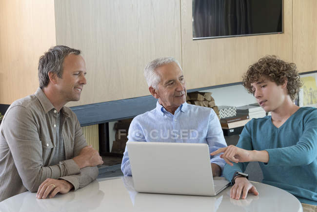 Feliz hombre mayor con hijo y nieto sentado en la mesa con portátil en la sala de estar - foto de stock