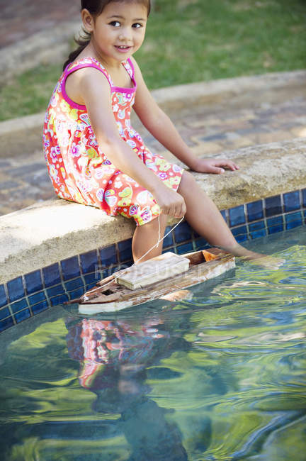 Menina sentada na borda da piscina com barco de brinquedo na água — Fotografia de Stock