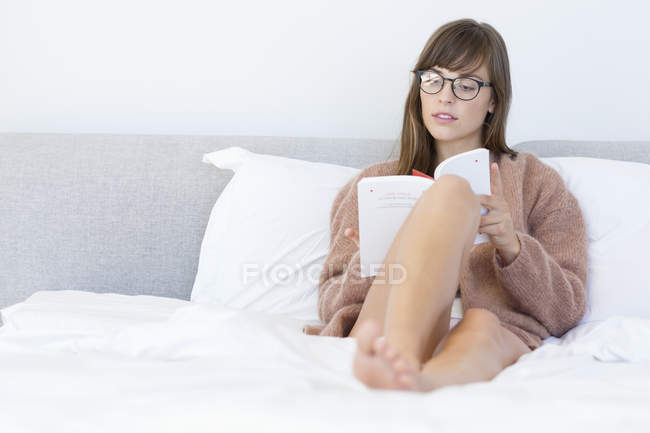 Focused young woman sitting on bed and reading book — Stock Photo