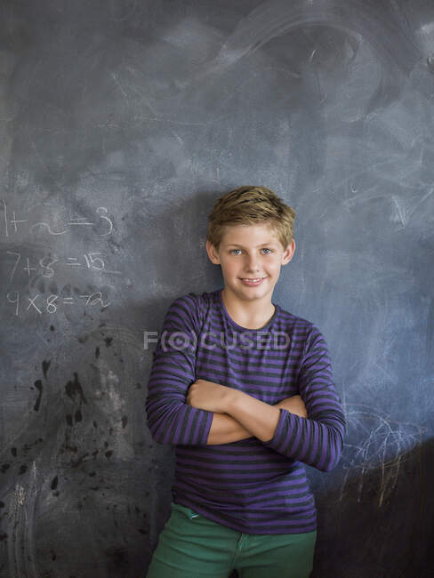 Garçon souriant les bras croisés devant un tableau noir dans une salle de classe — Photo de stock