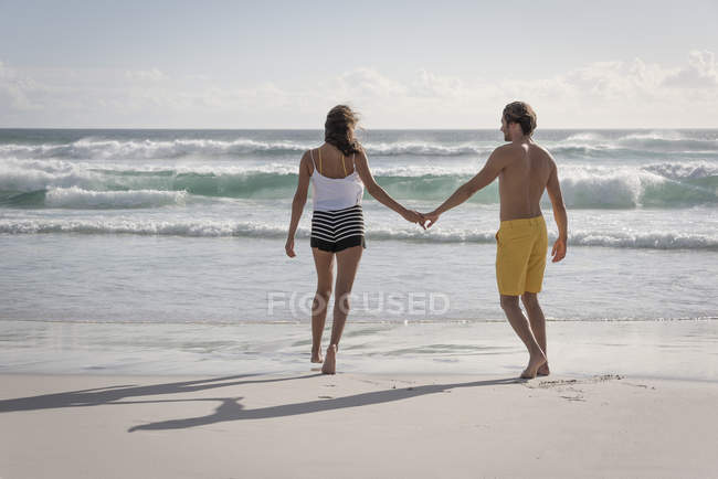 Rückansicht eines jungen Paares, das Händchen haltend am Strand spaziert — Stockfoto