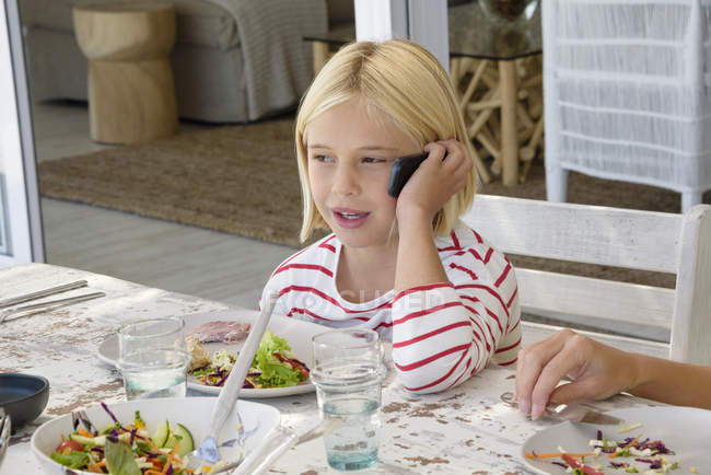 Little girl talking on mobile phone while eating meal — Stock Photo