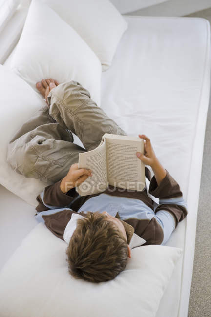 Menino deitado no sofá e livro de leitura — Fotografia de Stock