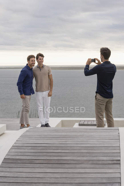 Man taking picture of friends with mobile phone on wooden terrace at lake — Stock Photo