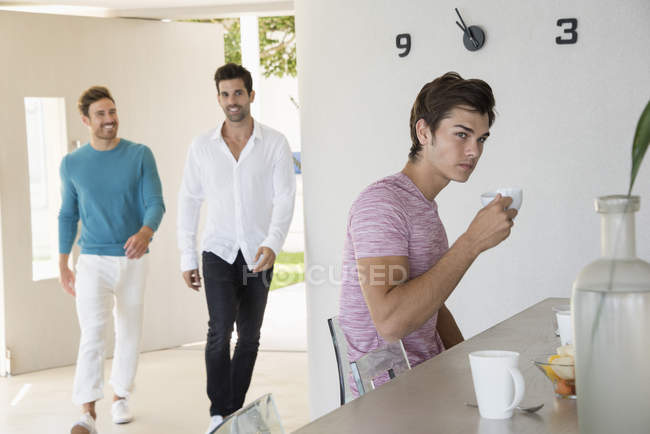 Jeune homme prenant un café à la maison avec des amis marchant sur le fond — Photo de stock