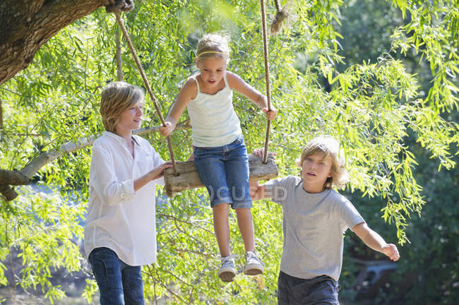 Lächelnde Geschwister spielen in Baumschaukel im Sommergarten — Stockfoto