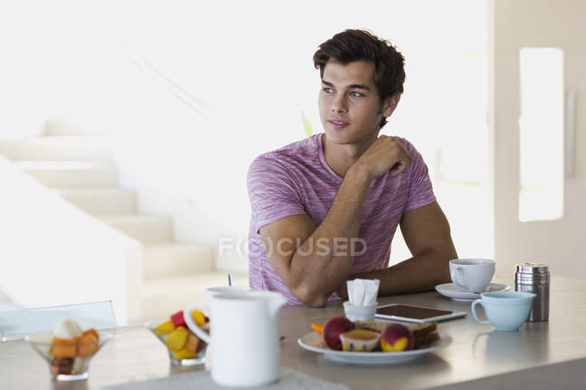 Молодой человек сидит за кухонным столом с чашкой кофе — стоковое фото