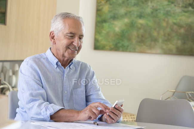 Homem sênior feliz usando telefone celular ao fazer papelada em casa — Fotografia de Stock