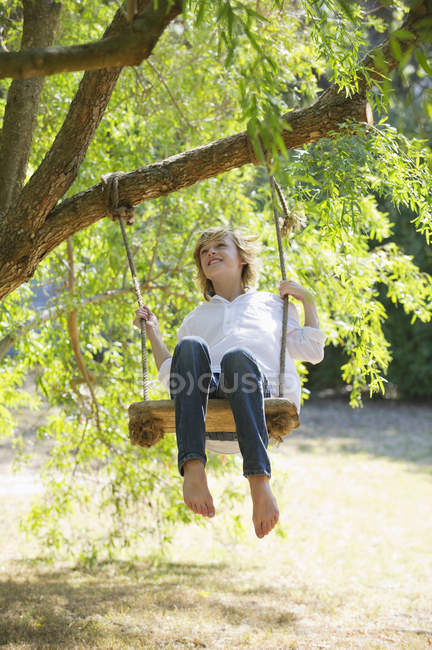 Happy barefoot teenage boy swinging on tree in summer countryside — Stock Photo