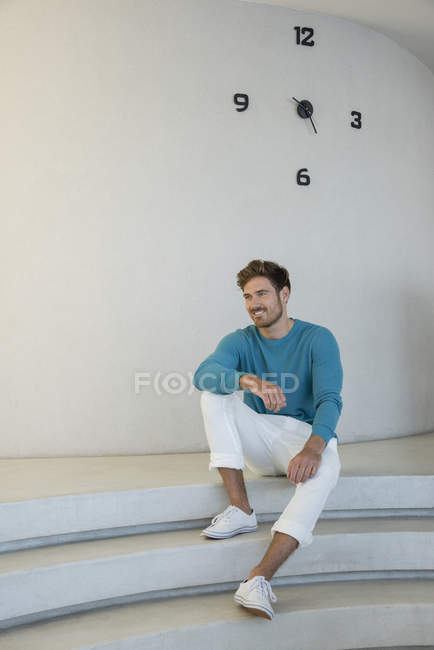 Glücklicher junger Mann sitzt auf Stufen vor weißer Wand mit Uhr — Stockfoto