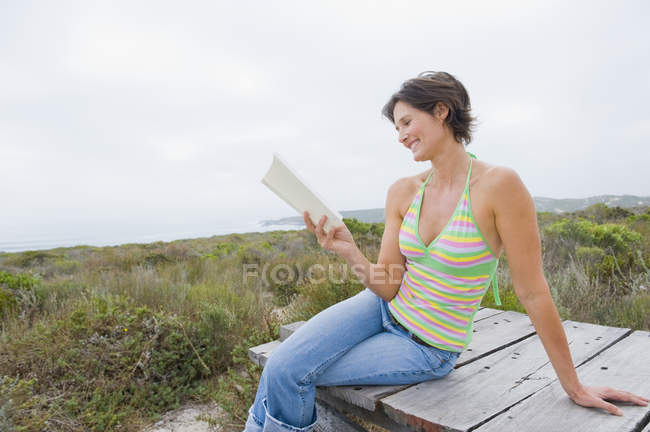 Sorrindo mulher sentada no calçadão na natureza e lendo um livro — Fotografia de Stock
