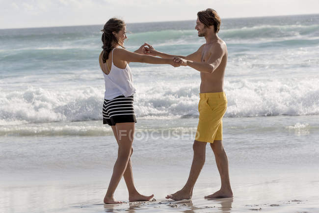 Jovem casal brincalhão de pé na praia de mãos dadas — Fotografia de Stock
